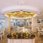 Elegant cruise ship restaurant with white chairs and a stained-glass ceiling centerpiece.