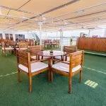 A spacious outdoor sun deck or rooftop bar on a cruise ship, featuring many sets of wooden tables and chairs with white cushions, a light fabric awning overhead, and a surface covered in artificial green grass.