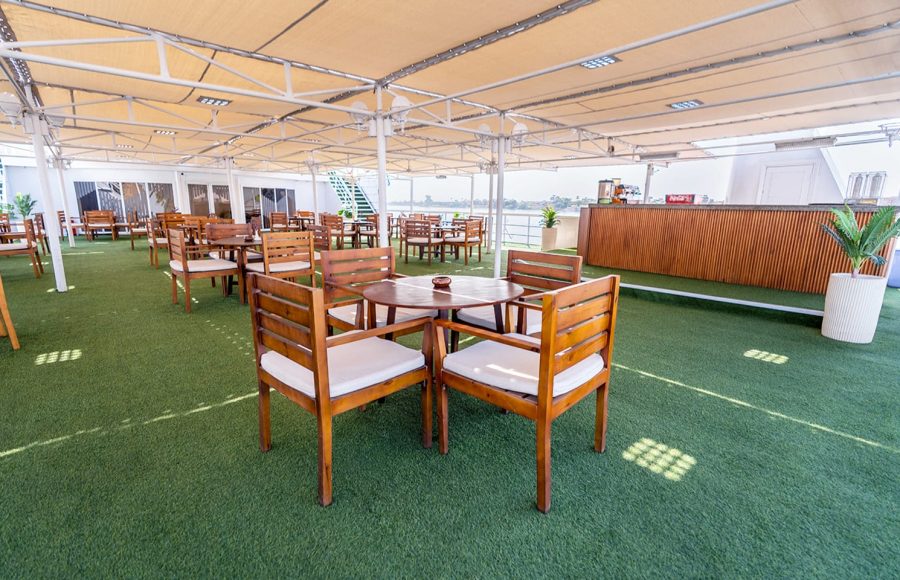 A spacious outdoor sun deck or rooftop bar on a cruise ship, featuring many sets of wooden tables and chairs with white cushions, a light fabric awning overhead, and a surface covered in artificial green grass.