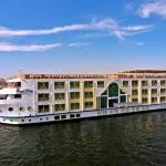 A large, white, multi-deck Nile cruise ship named "Royal Viking" sailing on the water under a clear blue sky, featuring distinctive green-framed windows and a central arched entrance area on the side.