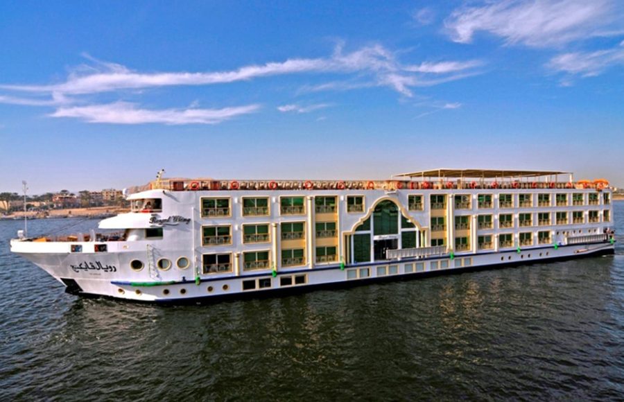 A large, white, multi-deck Nile cruise ship named "Royal Viking" sailing on the water under a clear blue sky, featuring distinctive green-framed windows and a central arched entrance area on the side.