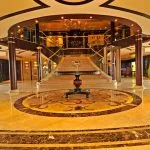 A lavish cruise ship lobby with highly polished marble floors, a grand central staircase with dark wood railings leading to a mezzanine, and a round central table decorated with a single glass.