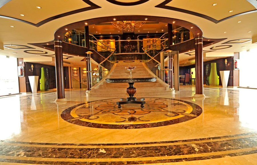 A lavish cruise ship lobby with highly polished marble floors, a grand central staircase with dark wood railings leading to a mezzanine, and a round central table decorated with a single glass.