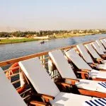 A row of wooden sun loungers with white cushions and blue and white striped towels lined up along the railing of a cruise ship deck, facing a wide, scenic view of the Nile River and the desert banks in the distance.