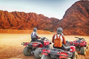 Two Travelers on red quad bikes looking out at a rugged, reddish desert landscape with mountains in the background, typical of a safari excursion from Safaga Port.