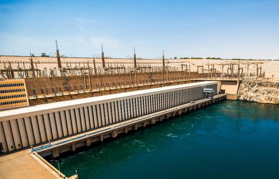 An aerial side view of the Aswan High Dam, showing its massive concrete structure, turbines, and the reservoir's blue water.