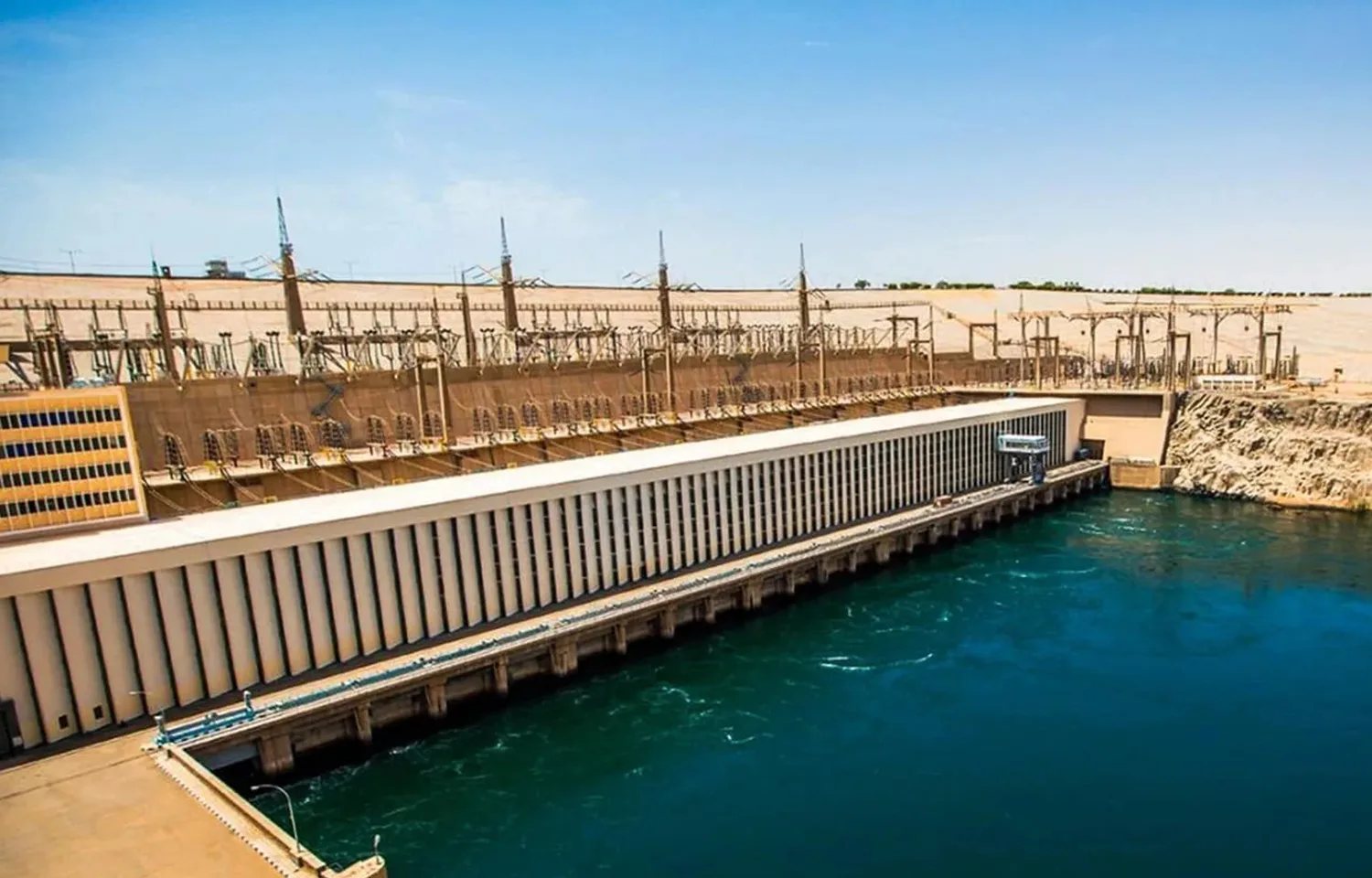 An aerial side view of the Aswan High Dam, showing its massive concrete structure, turbines, and the reservoir's blue water.
