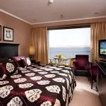 Interior of a cruise cabin looking out through a large window toward a private balcony and the sea.
