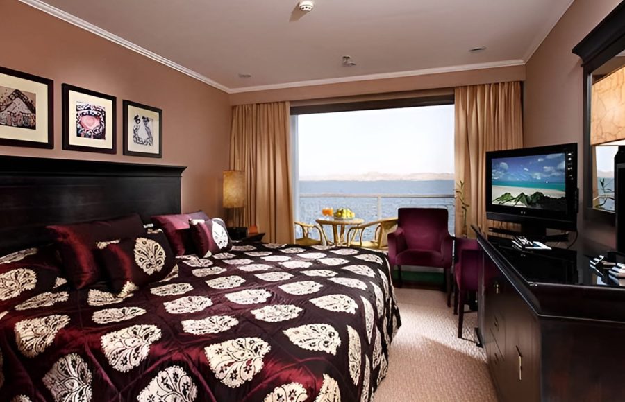 Interior of a cruise cabin looking out through a large window toward a private balcony and the sea.