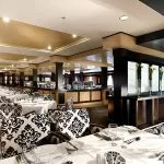 A large cruise ship dining hall with tables set with white linens and chairs with black and white patterned upholstery.