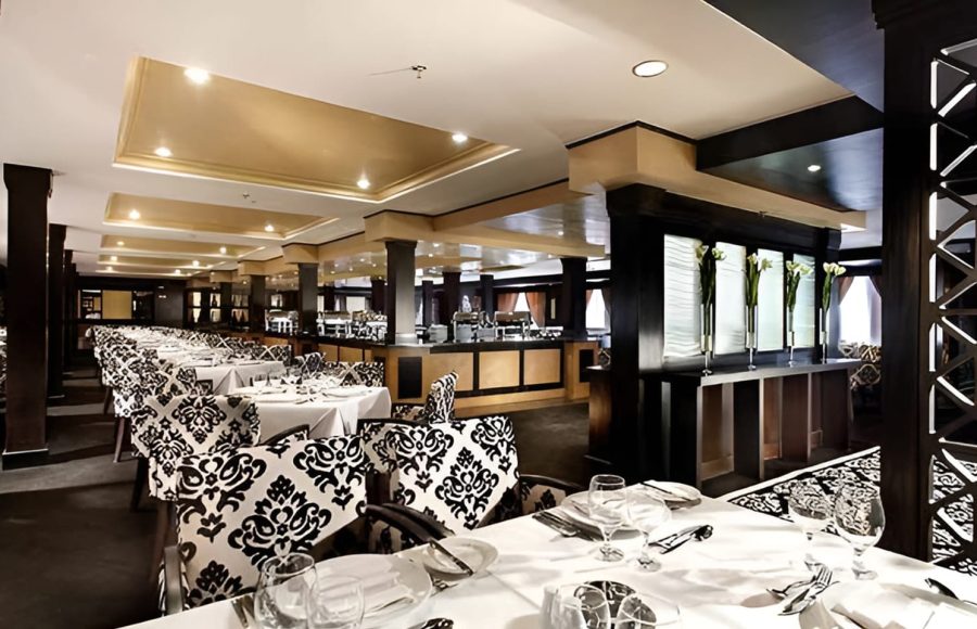 A large cruise ship dining hall with tables set with white linens and chairs with black and white patterned upholstery.