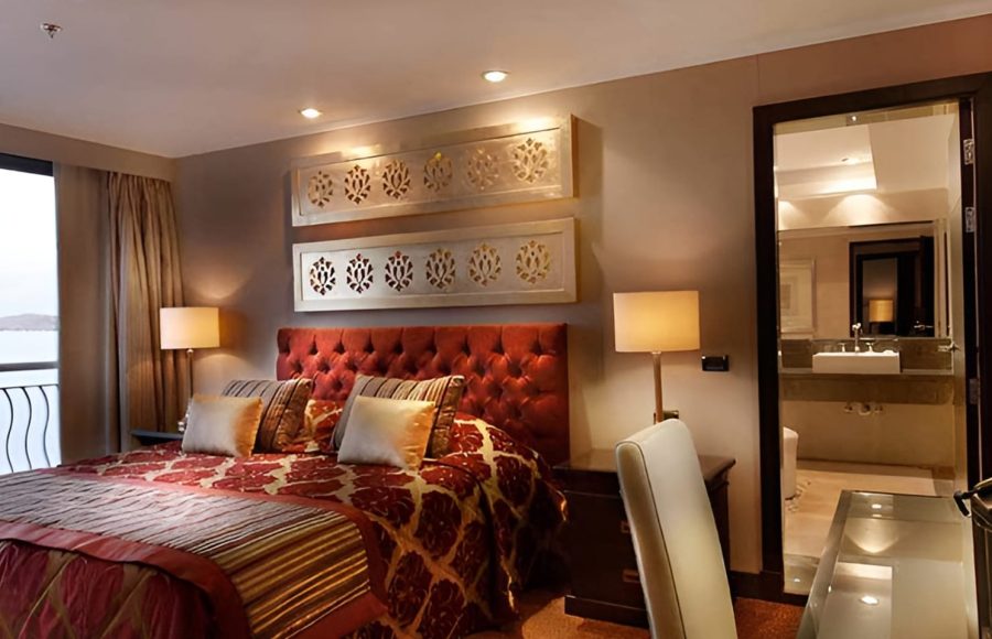 A luxurious cruise ship cabin with a large double bed featuring a red tufted headboard and gold-patterned bedding.