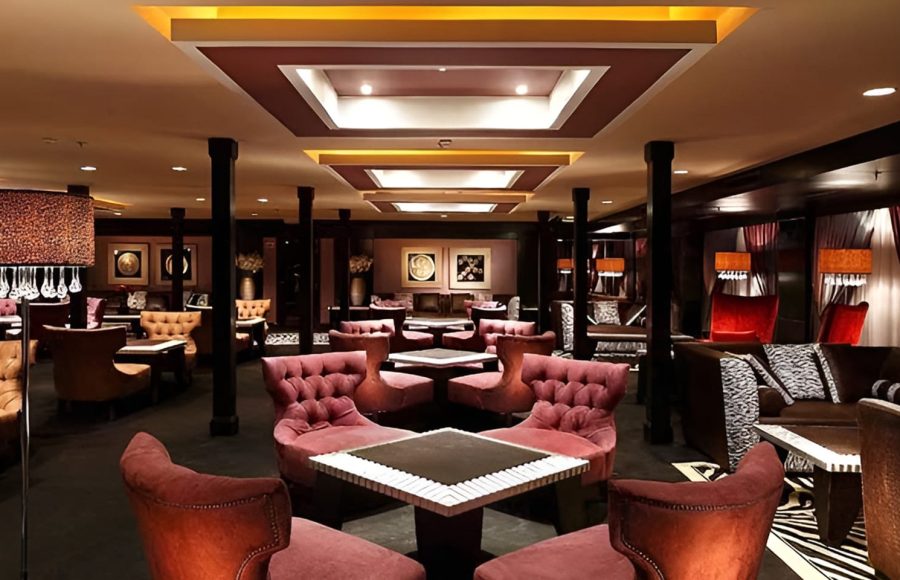 A wide lounge area on a cruise ship with purple velvet tufted chairs and modern recessed ceiling lighting.