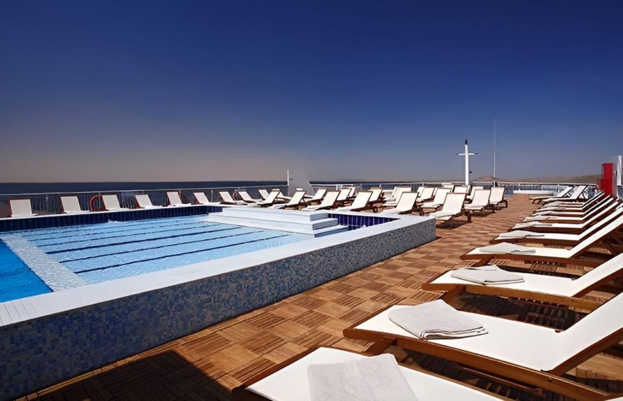 An outdoor ship deck with a swimming pool and several rows of wooden lounge chairs under a clear blue sky.