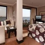 A large, luxurious ship suite divided into a living area with a sofa and armchairs and a sleeping area with a bed, both offering views of a private balcony and the water.
