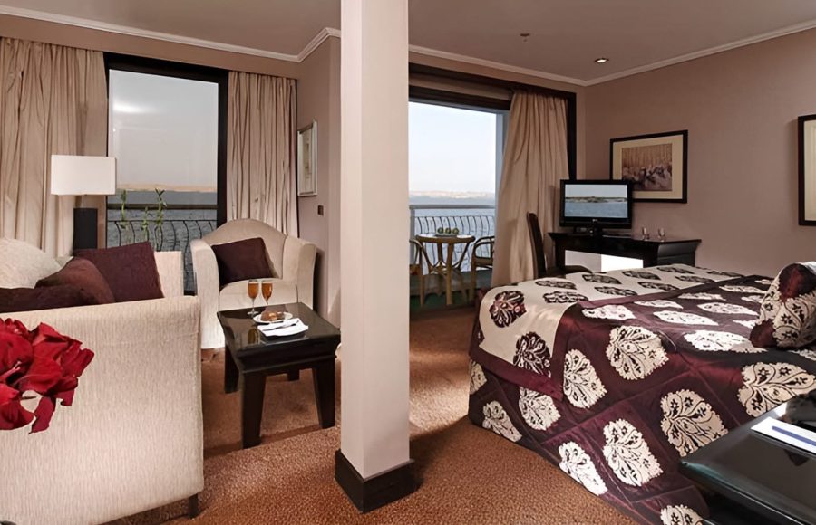 A large, luxurious ship suite divided into a living area with a sofa and armchairs and a sleeping area with a bed, both offering views of a private balcony and the water.