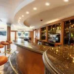 A curved, luxurious wooden bar with a dark, polished countertop and leather bar stools inside a cruise ship lounge.