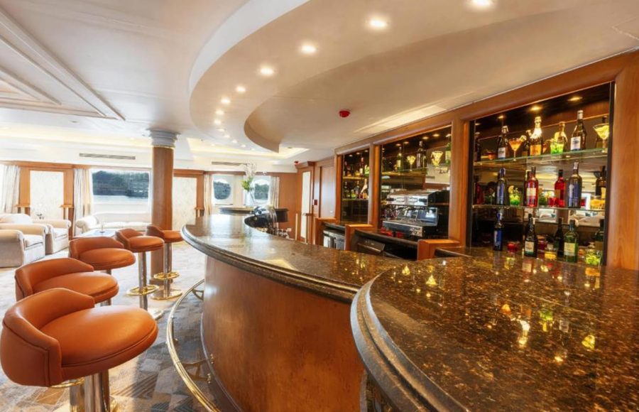 A curved, luxurious wooden bar with a dark, polished countertop and leather bar stools inside a cruise ship lounge.