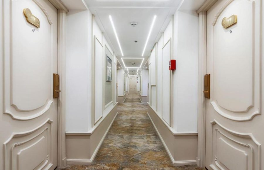 Long, symmetrical hallway of a cruise ship lined with light-colored cabin doors and illuminated by strip lighting.
