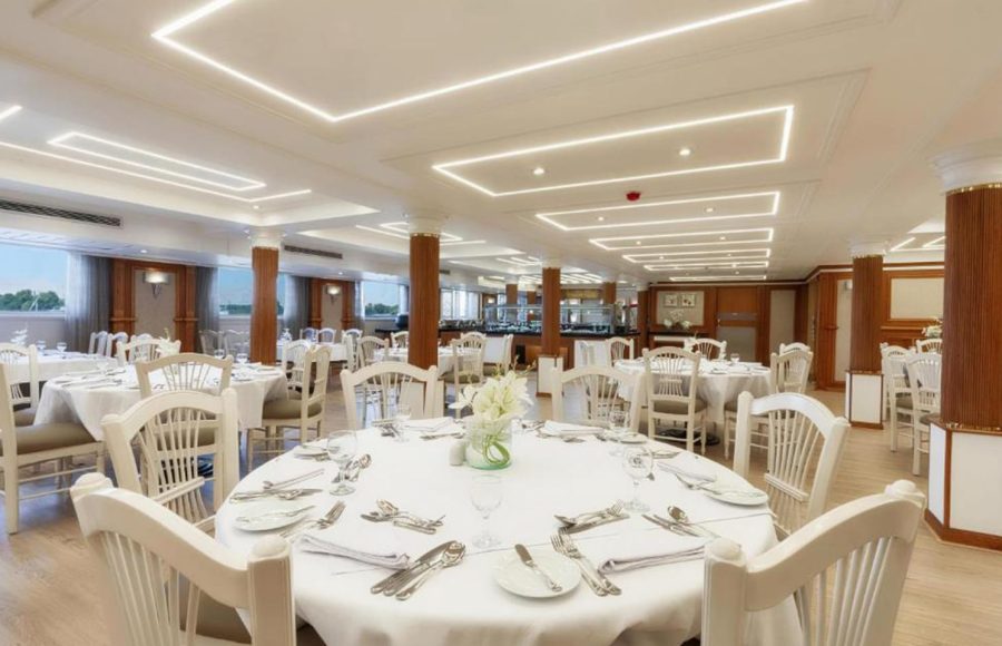 Bright, elegant cruise ship dining room with white-clothed round tables and white wooden chairs.