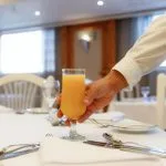 A waiter's hand placing a tall glass of orange juice on a white-clothed, set dining table in a cruise ship restaurant.