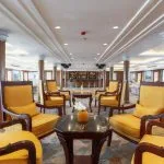 A wide view of a cruise ship dining room with multiple white-clothed round tables and white wooden chairs.