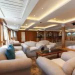 Interior view of a cruise ship lobby lounge with multiple beige couches and a dark wooden bar in the background.