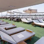 The sunny sundeck of a cruise ship, featuring multiple rows of wooden loungers with white cushions and orange towels, set on green artificial grass, under a large fabric awning, with a view of the river and distant shoreline.