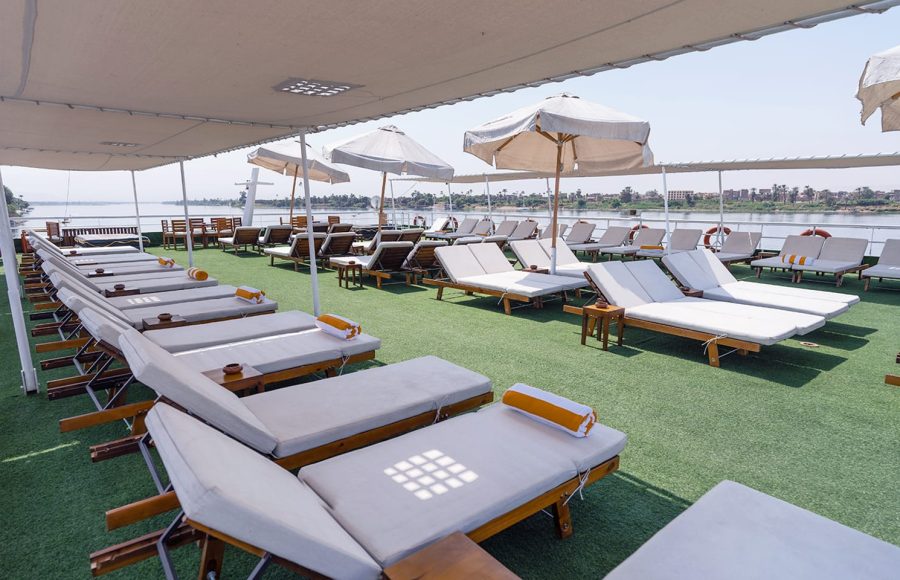 The sunny sundeck of a cruise ship, featuring multiple rows of wooden loungers with white cushions and orange towels, set on green artificial grass, under a large fabric awning, with a view of the river and distant shoreline.