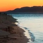 A beautiful sunrise in sharm el sheikh egypt over a beach lined with straw umbrellas and lounge chairs, showcasing the serene Red Sea and distant mountains, perfect for an Egypt tour cairo and red sea.