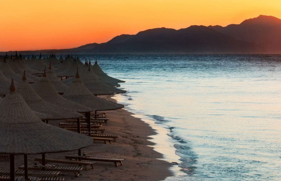 A beautiful sunrise in sharm el sheikh egypt over a beach lined with straw umbrellas and lounge chairs, showcasing the serene Red Sea and distant mountains, perfect for an Egypt tour cairo and red sea.