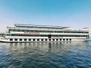 Nile Symphony cruise ship sailing on the river under a clear blue sky. Side view of the white and green Nile Symphony cruise boat on the water.
