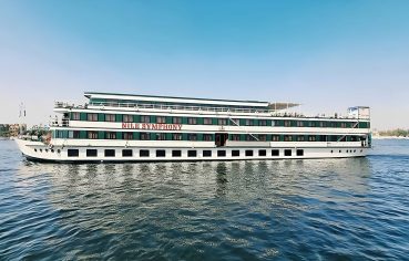 Nile Symphony cruise ship sailing on the river under a clear blue sky. Side view of the white and green Nile Symphony cruise boat on the water.