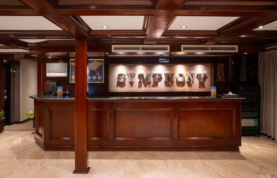 Ship's reception desk featuring a high counter made of dark wood and a backlit "SYMPHONY" sign against a patterned wall.