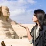 A young woman blowing a kiss towards the camera with the ancient Great Sphinx of Giza and a pyramid in her 6 days Cairo and Hurghada Holiday Package.